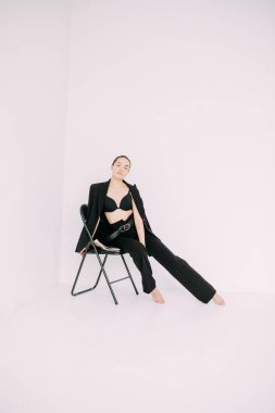 Siyah ceketli, pantolonlu ve sütyenli genç esmer kadın sandalyede çıplak ayakla oturuyor. Beyaz arkaplan izole görüntü.