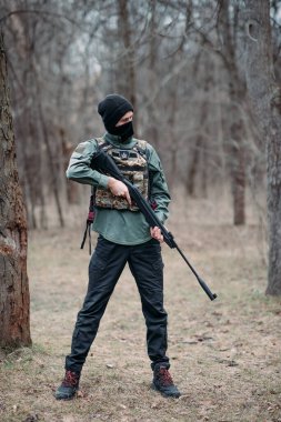 Genç adam elinde pompalı tüfekle ayakta duruyor. Ormanda çelik yelek ve kar maskesi giyiyor..