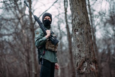 Genç adam elinde pompalı tüfekle ayakta duruyor. Ormanda çelik yelek ve kar maskesi giyiyor..