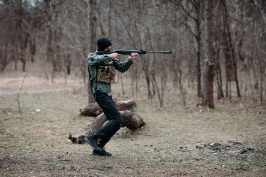 Delikanlı ormanda çelik yelek ve kar maskesi içinde pompalı tüfekle pratik yapıyor ve nişan alıyor..