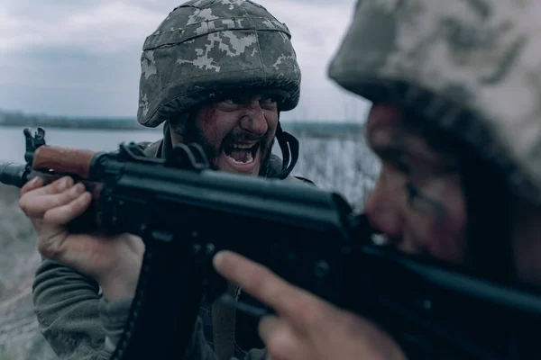 2人のウクライナ兵が空と川の背景に対してロシアの敵にカラシニコフのアサルトライフルから叫び ウクライナにおけるロシアの軍事侵攻の概念 ウクライナとヨーロッパでの戦争 — ストック写真