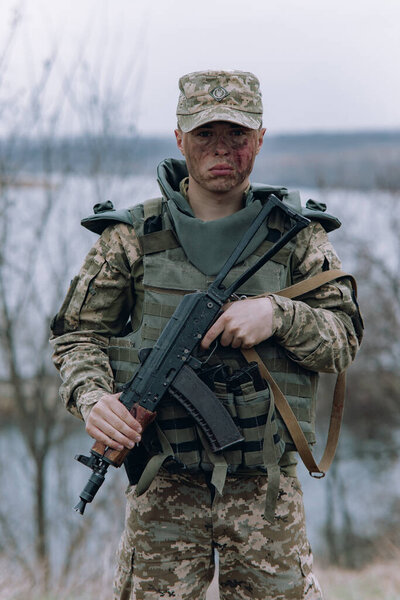 Украинский солдат стоит с автоматом Калашникова в руках на фоне неба и реки. Концепция военного вторжения России на Украину. Война на Украине и в Европе.