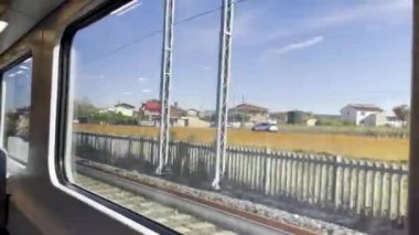Trenin penceresinden manzaraya köy evleri, arabalı yollar, çitler ve mavi gökyüzü.