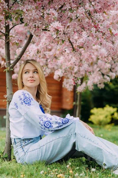 年轻的金发女子穿着乌克兰语绣花衬衫躺在草坪上 靠近盛开的樱花树 — 图库照片