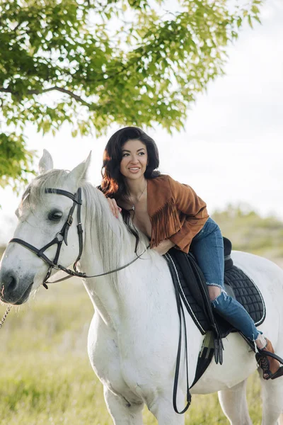 Young woman in unbuttoned jacket sits horseback on white horse on meadow.