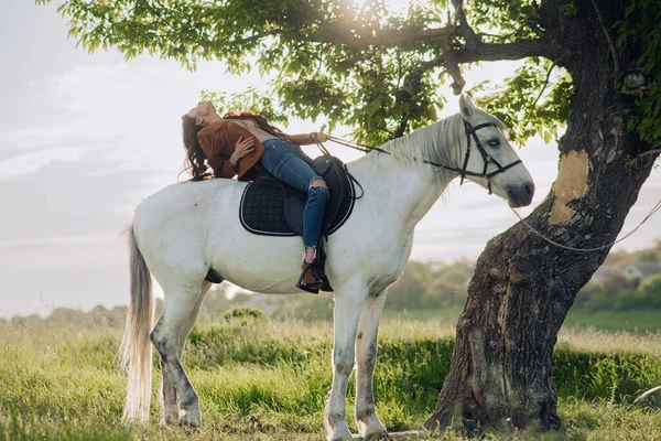 Young woman in unbuttoned jacket sits horseback on white horse near tree and enjoys by walk on meadow.
