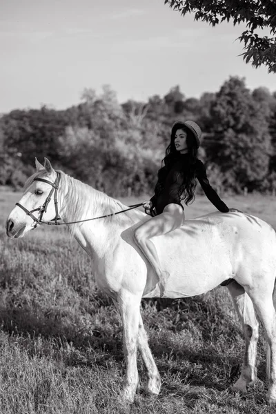 Young woman in bodysuit and hat sits horseback on white horse on meadow. Black and white image.