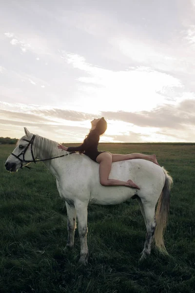 Young woman in bodysuit sits horseback on white horse on meadow at sunset.