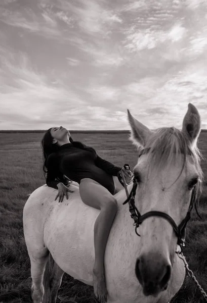 Young dreamy woman in bodysuit sits horseback on white horse on meadow at sunset. Black and white image.