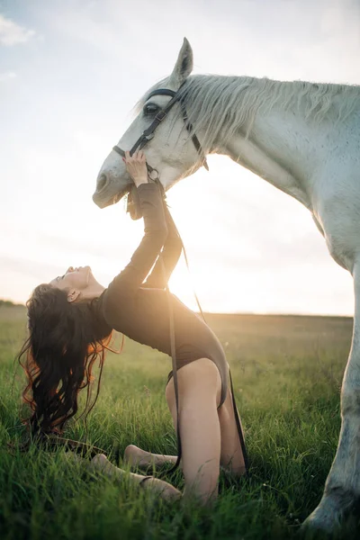 若いです夢のような女性ひざまずいて草の上にボディスーツとホールド白い馬によって花嫁に対して牧草地と空の背景で日没 — ストック写真