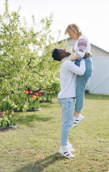 Межрасовая пара веселится и смеется на лужайке в весеннем саду, одетые в украинские традиционные этнические вышитые рубашки. Понятие любовных отношений и единства между различными человеческими расами.