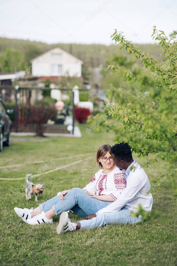 Interracial couple sits on grass in spring garden dressed in Ukrainian traditional ethnic embroidered shirts. Concept of love relationships and unity between different human races.