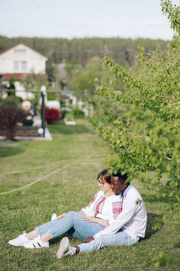 Interracial couple sits on grass in spring garden dressed in Ukrainian traditional ethnic embroidered shirts. Concept of love relationships and unity between different human races.