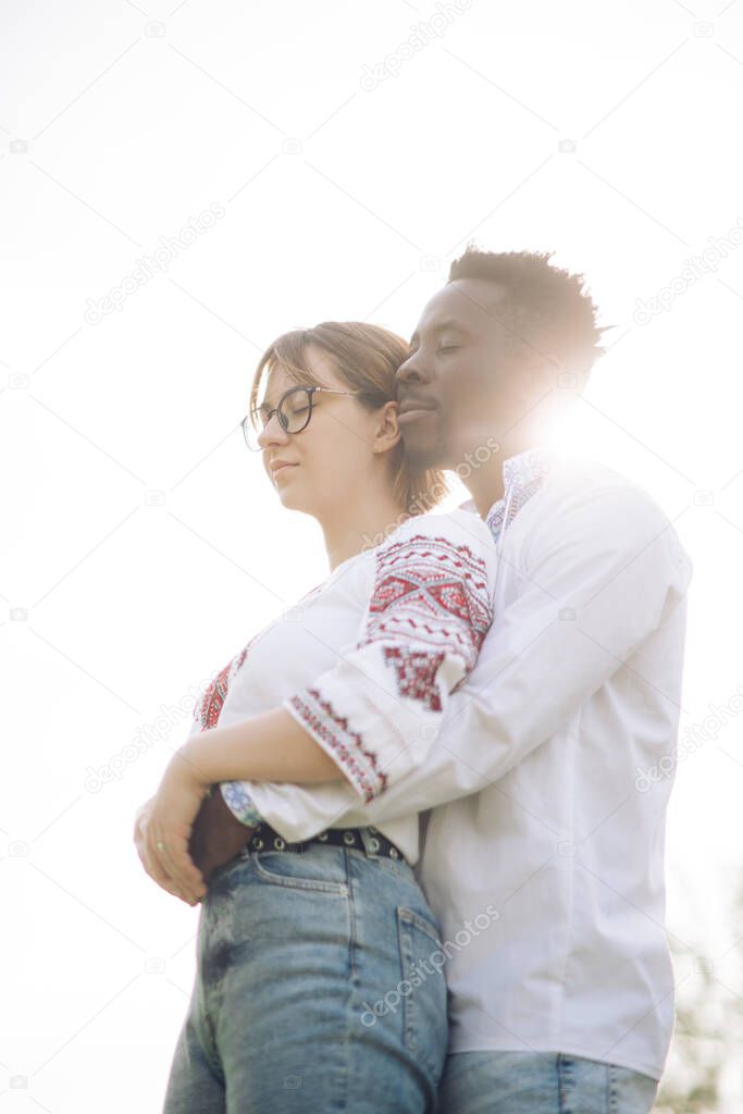 Interracial couple embraces against sky background dressed in Ukrainian traditional ethnic embroidered shirts. Concept of love relationships and unity between different human races. Backlight.