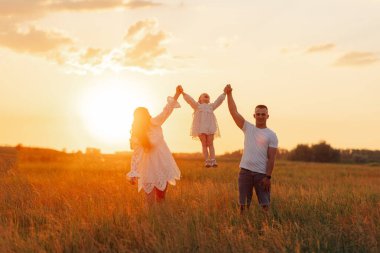 Çocuklu mutlu ebeveynler gün batımında gökyüzü arka planına karşı çayırda kızlarının elleriyle oynayıp kaldırıyorlar..