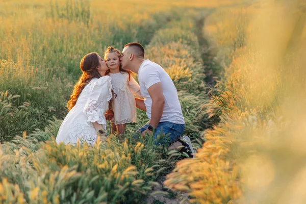 Happy parents kiss their daughter and play with her among meadow at sunset.