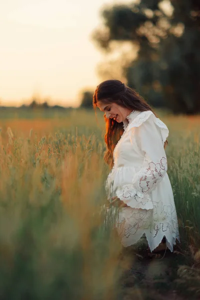 若いです幸せな妊娠中の女性は日没時に牧草地の間で白いドレスを歩く — ストック写真
