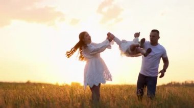 Mutlu genç ebeveynler gün batımında çayırda gezerken eğleniyor, oynuyor, ellerinden kaldırıyor ve kızlarını havaya fırlatıyor..