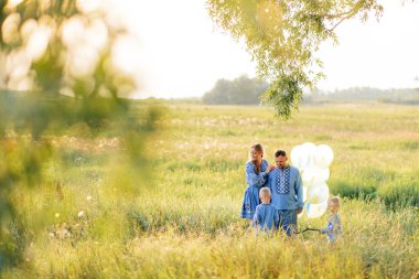 İki ikiz oğlu olan mutlu ebeveynler ellerinde hava balonlarıyla çayırlarda yürüyorlar. Ukrayna ulusal nakışlı gömlekler giyiyorlar..