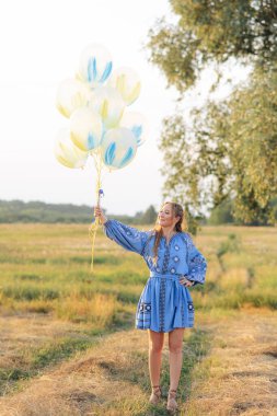 Mutlu Ukraynalı kadın elinde balonlarla çayırda yürüyor. Ukrayna ulusal nakışlı bir elbise giyiyor..