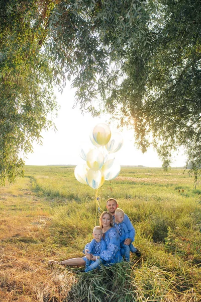 Happy parents with two their twins sons sit on grass and rest on meadow with air balloons in their hands. They are dressed in Ukrainian national embroidered shirts.