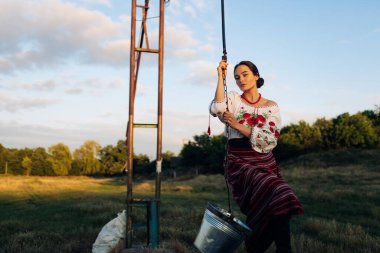 Young Ukrainian woman stands in traditional national embroidered shirt and skirt and draws water from well on meadow at sunset. Ethnic ukrainian national clothes style, embroidered shirt. Rural scene.