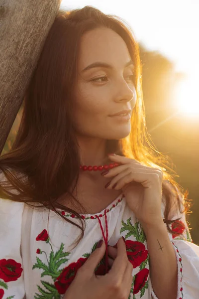 Portrait of young Ukrainian woman in traditional national embroidered shirt and necklace near wooden fence on meadow at sunset. Ethnic ukrainian national clothes style, embroidered shirt.