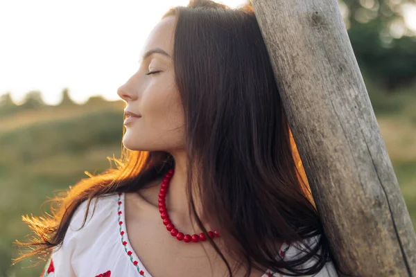 Portrait of young Ukrainian woman in traditional national embroidered shirt and necklace near wooden fence on meadow at sunset. Ethnic ukrainian national clothes style, embroidered shirt.