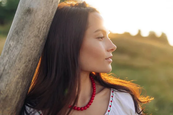 Portrait of young Ukrainian woman in traditional national embroidered shirt and necklace near wooden fence on meadow at sunset. Ethnic ukrainian national clothes style, embroidered shirt.