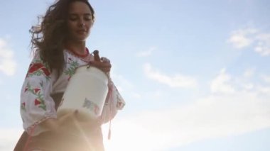 Ukrainian woman stands in traditional national embroidered shirt and skirt and pours out water from can against sky background. Ethnic ukrainian national clothes style, embroidered shirt.