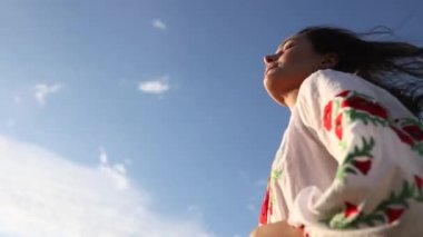 Young Ukrainian woman stands and corrects her hair in traditional national embroidered shirt against sky background. Ethnic ukrainian national clothes style, embroidered shirt. Copy space.