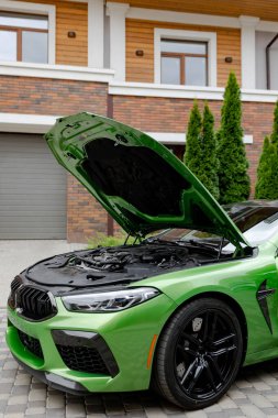 Modern green car with opened bonnet is on parking near garage gate of house.