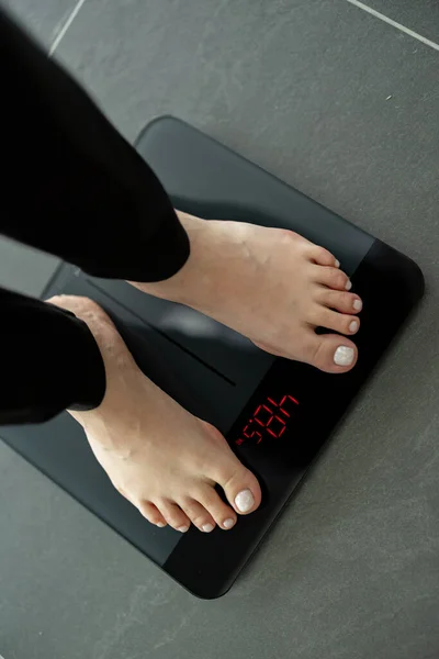 Woman measures her weight on floor scales. Female feet are on scales against the background of floor. The concept of diet and healthy lifestyle. Top view to visible numbers.