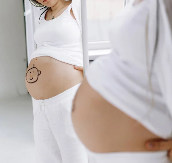 Pregnant woman looks on her reflection in mirror with drawn smiling baby face on her belly . Concept of love and care during pregnancy, expecting and born baby.