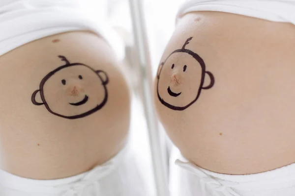 Pregnant woman looks on her reflection in mirror with drawn smiling baby face on her belly . Concept of love and care during pregnancy, expecting and born baby.