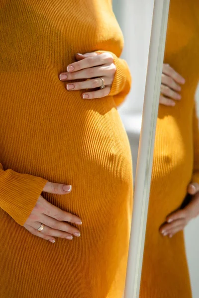 Pregnant woman in dress looks on her reflection in mirror. Concept of love and care during pregnancy, expecting and born baby.