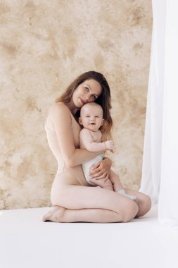 Genç anne beyaz perdelerin yanında oturuyor ve bebeğini kucaklıyor..