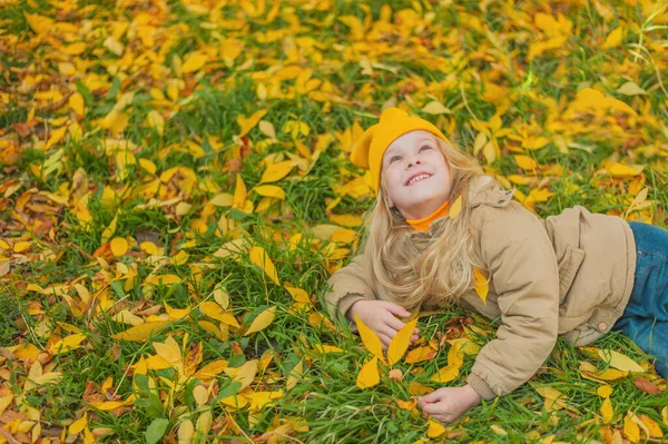 Una niña se acuesta en un césped cubierto de hojas amarillas de otoño y mira hacia arriba con una sonrisa en su cara La niña está feliz con el cálido y soleado otoño colorido —  Fotos de Stock