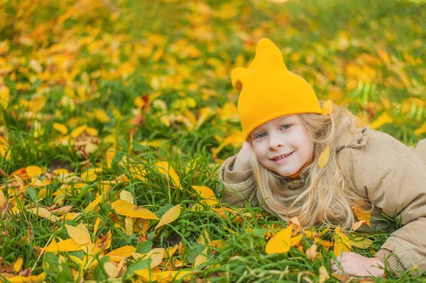 Usmívající se školačka s blond vlasy ve žlutém klobouku leží na trávě s podzimními listy portrét usměvavé krásné holčičky s dlouhými blond vlasy na podzim. Dívka leží na trávníku — Stock fotografie