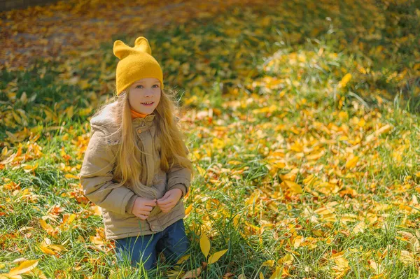 Belle enfant souriante en automne par beau temps s'assoit sur l'herbe avec des feuilles jaunes petite fille préscolaire blonde aux cheveux longs dans un chapeau jaune sur le fond d'une pelouse avec des feuilles d'automne jaunes — Photo
