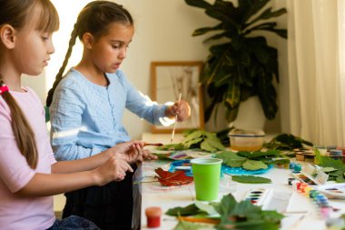 Kızlar sonbahar yapraklarını boyarlar. Gouache, fırça ve ahşap bir masada çeşitli sonbahar yaprakları. Çocuk sanat projesi. Kuru sonbahar yapraklarında renkli elle boyanmış.