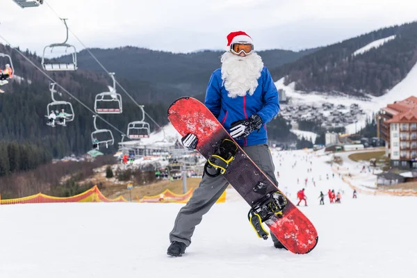 Un homme en chapeau de Père Noël avec un snowboard dans une station de ski — Photo