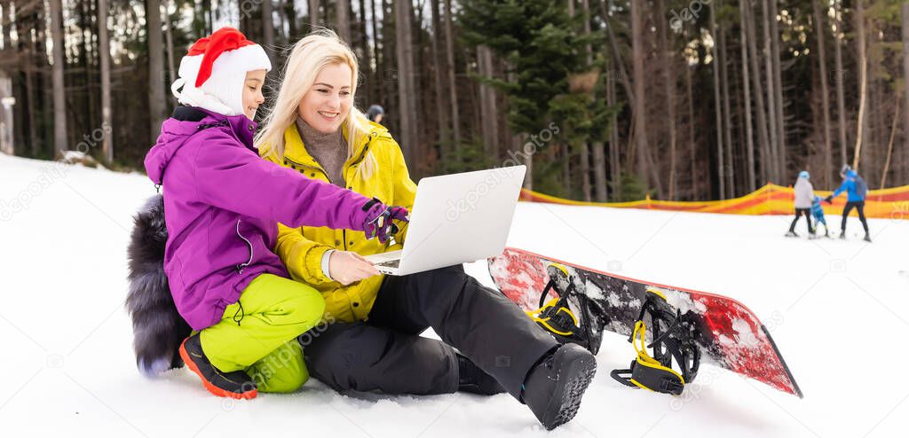 mother and daughter with a snowboard using laptop on snowy mountain