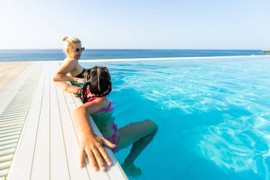 Bikinili genç anne ve kız havuz kenarında dinlenirken eğleniyorlar. Yaz tatilinde mutlu bir aile. Anne ve bebek havuzun kenarında oturuyorlar. Macera, dinlenme ve dinlenme konsepti.