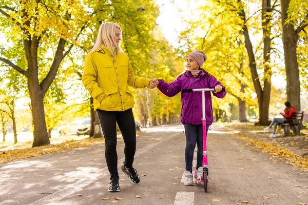 엄마와 딸은 가을에 공원을 걷고 있습니다. 여자 애가 스쿠터타고 있어요 — 스톡 사진