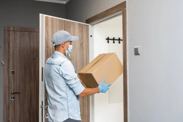Delivery. Cheerful young courier holding a cardboard box while standing at the entrance of apartment — Stock Photo, Image