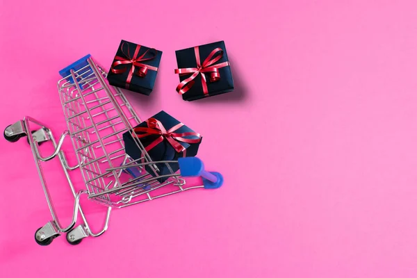 Δημιουργική ιδέα με τρόλεϊ αγορών με δώρα που απομονώνονται σε ροζ φόντο. Δώρα τυλιγμένα σε χαρτί κραφτ με κόκκινη κορδέλα και φιόγκο. — Φωτογραφία Αρχείου
