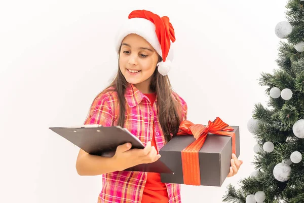 Sonriente niña divertida con sombrero rojo de Santa. Llevando un regalo de Navidad en la mano. Concepto de Navidad. sobre fondo blanco —  Fotos de Stock
