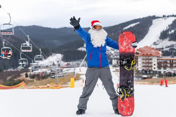 Egy férfi télapó kalapban, snowboarddal a síközpontban. — Stock Fotó