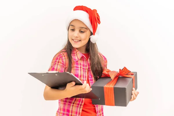 Sonriente niña divertida con sombrero rojo de Santa. Llevando un regalo de Navidad en la mano. Concepto de Navidad. sobre fondo blanco —  Fotos de Stock